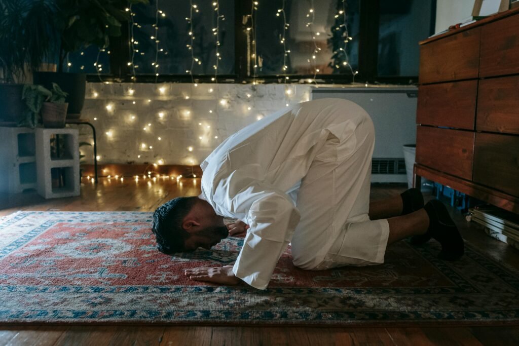 A Muslim man in traditional attire praying on a rug indoors with ambient lighting.