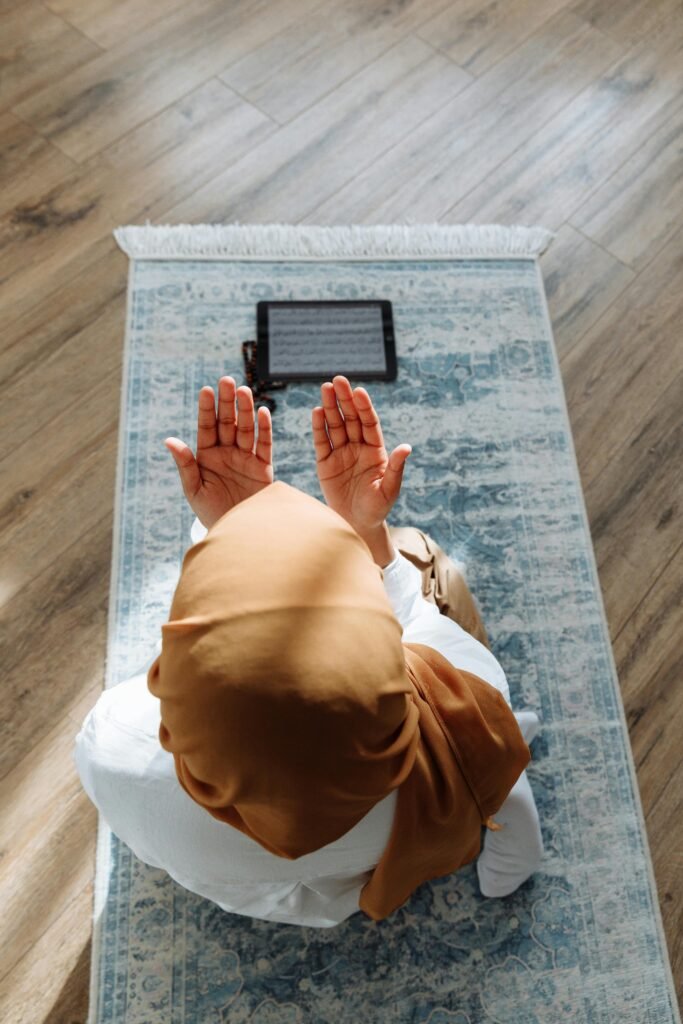 pexels-photo-7956746-7956746 A woman in a hijab prays on a rug with a tablet nearby, in a peaceful indoor setting.