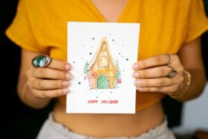 Woman holding a handmade holiday card with a gingerbread house illustration.