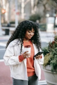 Cheerful young woman with curly hair using smartphone and holding coffee to go outdoors.